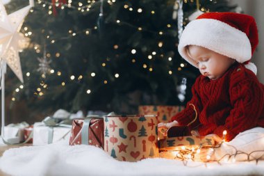 Noel Baba şapkalı sevimli bebek modern dekore edilmiş noel ağacının altında süslü hediye kutuları açıyor. Bebeğin ilk Noel 'i. Atmosferik tatil zamanı, Mutlu Noeller!