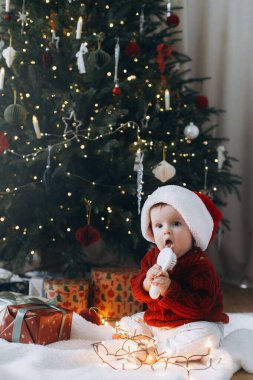 Noel Baba şapkalı sevimli bebek modern Noel ağacının altında çelenk ışıklarıyla şık bir şekilde duruyor. Atmosferik tatil zamanı, Mutlu Noeller!