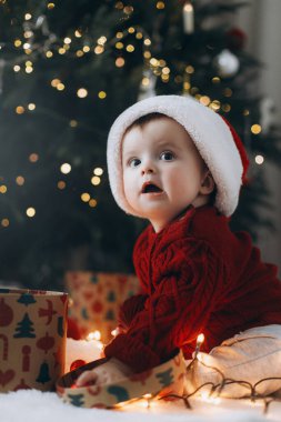 Noel Baba şapkalı sevimli bebek modern dekore edilmiş noel ağacının altında süslü hediye kutuları açıyor. Bebeğin ilk Noel 'i. Atmosferik tatil zamanı, Mutlu Noeller!