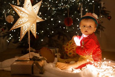 Noel Elf kostümlü sevimli bebek Noel arifesinde süslemeli Noel ağacının altında sihirli ışıkları olan hediye kutusuyla oturuyor. Bebeğin ilk Noel 'i. Atmosferik tatil zamanı, Mutlu Noeller!