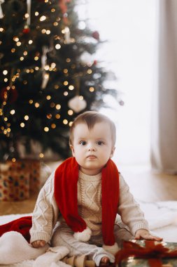 Süslü süveter giymiş sevimli mutlu bir bebek arka planda ışıklarla süslenmiş Noel ağacının üzerinde şık hediyelerle oturuyor. Atmosferik tatil zamanı, Mutlu Noeller!