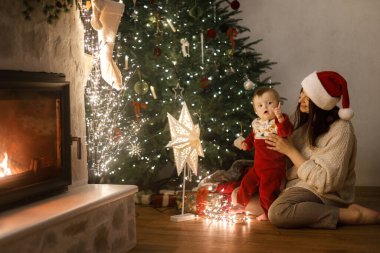 Şirin bebek ve anne rahat süveterler içinde şık bir Noel ağacının altında hediyeler ve şenlikli bir akşamda parlayan ışıklarla oturuyor. Mutlu Noeller ve mutlu tatiller! Aile sihirli xmas arifesinde