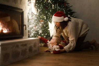 Şirin bebek ve anne rahat kazaklar içinde şık bir Noel ağacında hediyeler ve şenlikli bir akşamda parlayan ışıklarla emekliyor. Mutlu Noeller ve mutlu tatiller! Aile sihirli xmas arifesinde