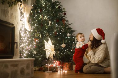 Şirin bebek ve anne rahat süveterler içinde şık bir Noel ağacının altında hediyeler ve şenlikli bir akşamda parlayan ışıklarla oturuyor. Mutlu Noeller ve mutlu tatiller! Aile sihirli xmas arifesinde