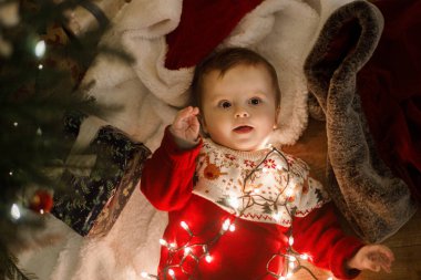 Sihirli Noel arifesinde şirin bir bebek. Mutlu Noeller ve mutlu tatiller! Parıldayan ışıklarla oynayan ve hediyelerle süslü Noel ağacının altında uzanan neşeli kazaklı mutlu çocuk.