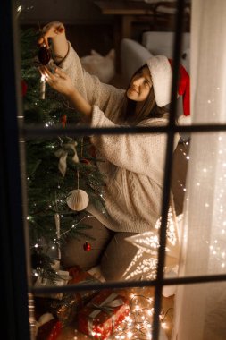 Noel Baba şapkalı, rahat süveterli, süslü Noel ağacını süsleyen, akşamları ışık saçan, pencereden bakan mutlu bir kadın. Mutlu Noeller! Sihirli xmas arifesi