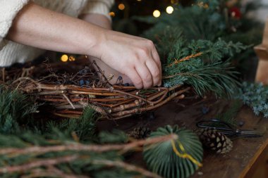 Noel çelengi yapıyorum. Eller çelengi köknar ve sedir dallarıyla düzenliyor, ve makas, kırsal yıldız ahşap masa üzerinde şık xmas ağacının arka planında. Kış tatili hazırlığı ve dekorasyonu