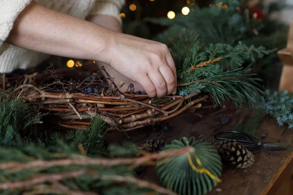 Noel çelengi yapıyorum. Eller çelengi köknar ve sedir dallarıyla düzenliyor, ve makas, kırsal yıldız ahşap masa üzerinde şık xmas ağacının arka planında. Kış tatili hazırlığı ve dekorasyonu