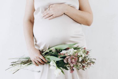 Güzel, hamile bir kadın kucağında çiçekleri yumuşak bir ışıkta tutuyor. Anneler Günün kutlu olsun! Sağlıklı hamilelik ve doğurganlık kavramı. Beyaz ipek elbiseli hamile kadın, hassas annelik.