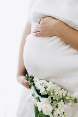 Güzel hamile kadın elinde çiçeklerle bebek şişliği tutuyor. Yaklaş. Anneler Günün kutlu olsun! Sağlıklı hamilelik ve doğurganlık kavramı. Beyaz ipek bornozlu hamile kadın, hassas annelik.