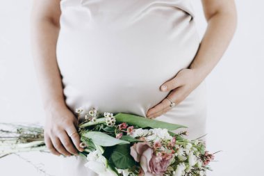 Elinde bebek şişliği ve güzel çiçekler olan şık hamile bir kadın. Sağlıklı hamilelik ve doğurganlık kavramı. Anneler Günün kutlu olsun! İpek elbiseli hamile kadın nazikçe sarılıyor göbeğine