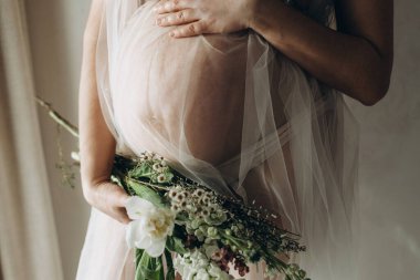 Güzel hamile bir kadın kucağında bebek şişliği, çiçekler ve yumuşak ışıkta poz veriyor. Sağlıklı hamilelik ve doğurganlık kavramı. Anneler Günün kutlu olsun! Peçeli hamile kadın nazikçe sarılıyor karnına