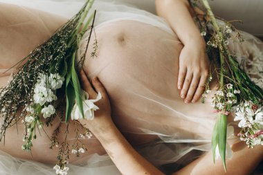 Elinde bebek şişliği ve güzel çiçekler olan şık hamile bir kadın. Sağlıklı hamilelik ve doğurganlık kavramı. Anneler Günün kutlu olsun! Hamile kadın peçenin altında uzanıyor ve yavaşça karnına sarılıyor.