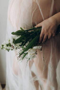 Güzel hamile bir kadın kucağında bebek şişliği, çiçekler ve yumuşak ışıkta poz veriyor. Sağlıklı hamilelik ve doğurganlık kavramı. Anneler Günün kutlu olsun! Peçeli hamile kadın nazikçe sarılıyor karnına