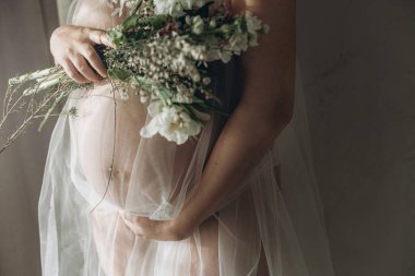Güzel hamile bir kadın kucağında bebek şişliği, çiçekler ve yumuşak ışıkta poz veriyor. Anneler Günün kutlu olsun! Sağlıklı hamilelik ve doğurganlık kavramı. Peçeli hamile kadın, hassas annelik.