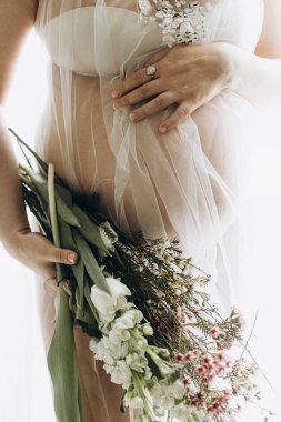 Güzel hamile bir kadın kucağında bebek şişliği, çiçekler ve yumuşak ışıkta poz veriyor. Sağlıklı hamilelik ve doğurganlık kavramı. Anneler Günün kutlu olsun! Peçeli hamile kadın nazikçe sarılıyor karnına