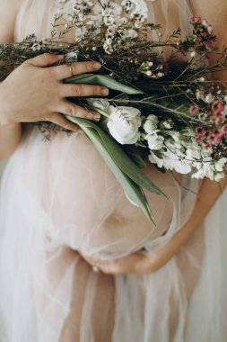 Elinde bebek şişliği ve güzel çiçekler olan şık hamile bir kadın. Sağlıklı hamilelik ve doğurganlık kavramı. Anneler Günün kutlu olsun! Peçeli hamile kadın poz veriyor ve nazikçe karnına sarılıyor.