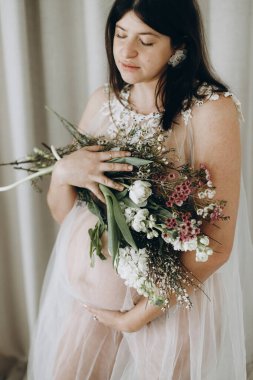 Elinde bebek şişliği ve güzel çiçekler olan şık hamile bir kadın. Sağlıklı hamilelik ve doğurganlık kavramı. Anneler Günün kutlu olsun! Peçeli hamile kadın poz veriyor ve nazikçe karnına sarılıyor.