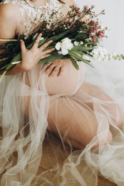 Elinde bebek şişliği ve güzel çiçekler olan şık hamile bir kadın. Sağlıklı hamilelik ve doğurganlık kavramı. Anneler Günün kutlu olsun! Peçeli hamile kadın poz veriyor ve nazikçe karnına sarılıyor.