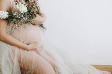 Elinde bebek şişliği ve güzel çiçekler olan şık hamile bir kadın. Sağlıklı hamilelik ve doğurganlık kavramı. Anneler Günün kutlu olsun! Peçeli hamile kadın poz veriyor ve nazikçe karnına sarılıyor.