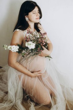 Elinde bebek şişliği ve güzel çiçekler olan şık hamile bir kadın. Sağlıklı hamilelik ve doğurganlık kavramı. Anneler Günün kutlu olsun! Peçeli hamile kadın poz veriyor ve nazikçe karnına sarılıyor.