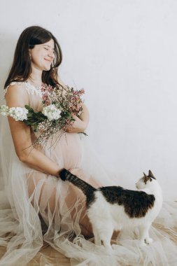 Mutlu hamile kadın elinde çiçeklerle kucağında tatlı kediyle oturuyor. Anneler Günün kutlu olsun! Sağlıklı hamilelik ve doğurganlık kavramı. Peçeli hamile kadın, hassas annelik.
