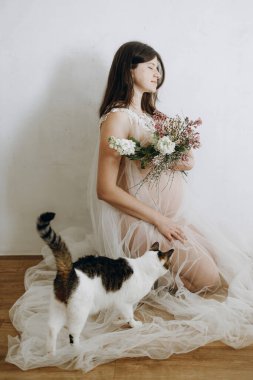 Havalı hamile kadın peçesinde güzel çiçeklerle bebek şişliği tutuyor ve kedisiyle oturuyor. Sağlıklı hamilelik ve doğurganlık kavramı. Anneler Günün kutlu olsun.!