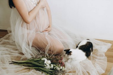 Havalı hamile kadın peçesinde güzel çiçeklerle bebek şişliği tutuyor ve kedisiyle oturuyor. Sağlıklı hamilelik ve doğurganlık kavramı. Anneler Günün kutlu olsun.!