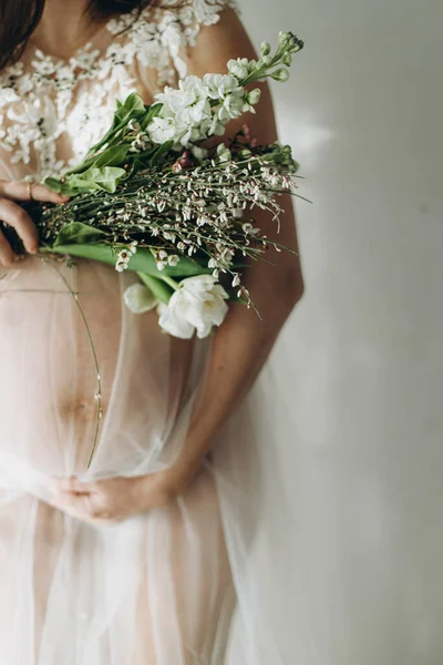 Güzel hamile bir kadın kucağında bebek şişliği, çiçekler ve yumuşak ışıkta poz veriyor. Sağlıklı hamilelik ve doğurganlık kavramı. Anneler Günün kutlu olsun! Peçeli hamile kadın nazikçe sarılıyor karnına