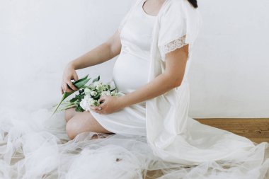 Güzel hamile kadın elinde çiçeklerle bebek şişliği tutuyor. Yaklaş. Anneler Günün kutlu olsun! Sağlıklı hamilelik ve doğurganlık kavramı. Beyaz ipek bornozlu hamile kadın, hassas annelik.
