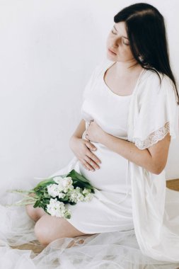 Elinde bebek şişliği ve güzel çiçekler olan şık hamile bir kadın. Sağlıklı hamilelik ve doğurganlık kavramı. Anneler Günün kutlu olsun! İpek bornozlu hamile kadın nazikçe sarılıyor karnına