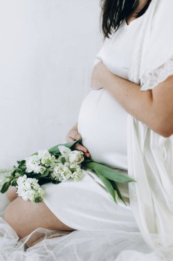 Elinde bebek şişliği ve güzel çiçekler olan şık hamile bir kadın. Sağlıklı hamilelik ve doğurganlık kavramı. Anneler Günün kutlu olsun! İpek bornozlu hamile kadın nazikçe sarılıyor karnına