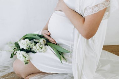 Elinde bebek şişliği ve güzel çiçekler olan şık hamile bir kadın. Sağlıklı hamilelik ve doğurganlık kavramı. Anneler Günün kutlu olsun! İpek bornozlu hamile kadın nazikçe sarılıyor karnına