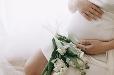 Güzel hamile kadın elinde çiçeklerle bebek şişliği tutuyor. Yaklaş. Anneler Günün kutlu olsun! Sağlıklı hamilelik ve doğurganlık kavramı. Beyaz ipek bornozlu hamile kadın, hassas annelik.