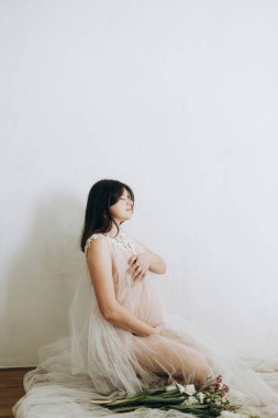 Elinde bebek şişliği ve güzel çiçekler olan şık hamile bir kadın. Sağlıklı hamilelik ve doğurganlık kavramı. Anneler Günün kutlu olsun! Peçeli hamile kadın poz veriyor ve nazikçe karnına sarılıyor.