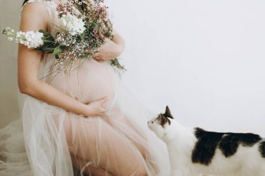 Havalı hamile kadın peçesinde güzel çiçeklerle bebek şişliği tutuyor ve kedisiyle oturuyor. Sağlıklı hamilelik ve doğurganlık kavramı. Anneler Günün kutlu olsun.!