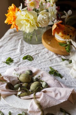 Mutlu Paskalyalar! Peçetedeki zarif yumurtalar, stanttaki paskalya pastası, bahar çiçekleri. Paskalya kutlaması masası, tatil durgun hayatı. Tebrik kartı ya da afiş