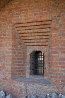 Eski bir manastırın duvarında küçük bir pencere. Pencere metal çubuklardan yapılmış bir kafes ile korunuyor. Kırmızı tuğla duvar.