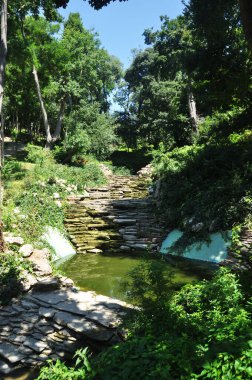 Parkta küçük bir gölet manzarası. Düz taşlardan oluşan bir göletin kıyısında. Yaz, güneşli gün.