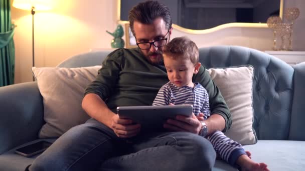 父とかわいい赤ちゃんの男の子は自宅でソファに座って一緒にデジタルタブレットで遊ぶ — ストック動画