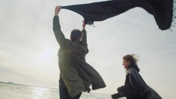 Una feliz pareja corriendo en la fría playa soleada en otoño — Vídeo de stock