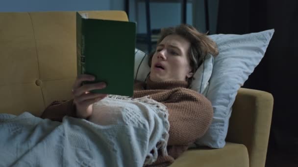 Cari tampan pria membaca buku berbaring di sofa di bawah selimut — Stok Video