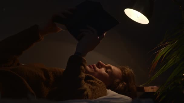 Красивий хлопець читає гучну книгу вночі, лежачи під лампою — стокове відео
