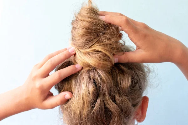 Meisje heft het misvormde broodje op haar hoofd met haar handen. Modern snel kapsel. Blauwe achtergrond. Blond krullend haar — Stockfoto