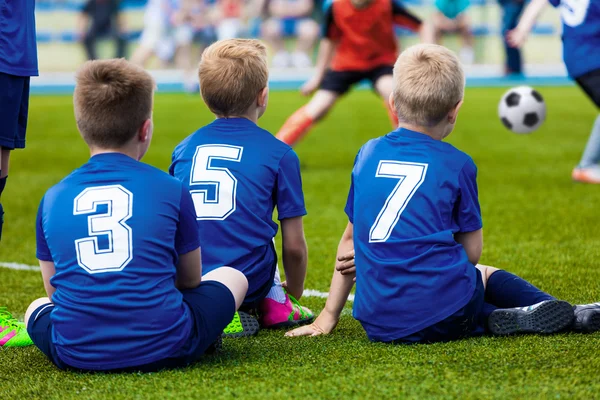 Bambini della giovane squadra di calcio. Ragazzi in divisa sportiva blu come giocatori di riserva seduti sul campo da calcio e a guardare la partita di calcio . — Foto Stock