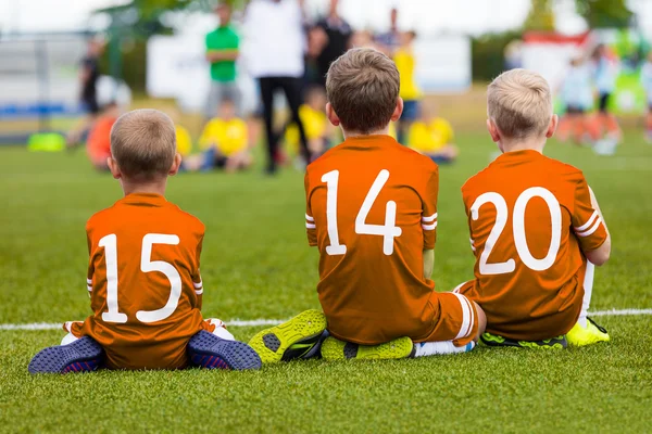 青少年足球橄榄球队的孩子们。男孩在橙色衬衫作为储备球员坐在足球场上和观看足球比赛. — 图库照片