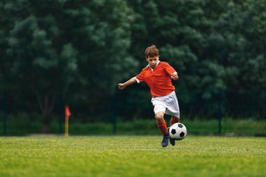 Futbol atışı. Çocuk çim tarlasında futbol topunu tekmeliyor. Genç futbolcu hareketli koşuyor, zıplıyor ve topa vuruyor. Genç seviye spor müsabakası. Kırmızı formalı futbolcu.