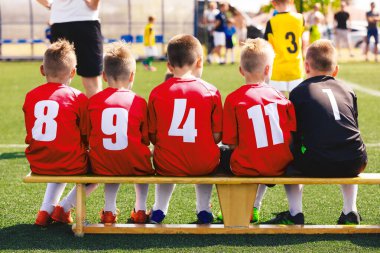 Kırmızı tişörtlü spor takımındaki çocuklar. Çocuklar yedek futbol bankında oturuyor. Okul futbol turnuvasındaki çocuklar. Arka planda futbol koçu ve yarışma oyunu