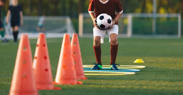 Close up boy running on football training pitch with ball in his hands. Player jumping over obstacles. Football training obstacle course. Children level soccer drill