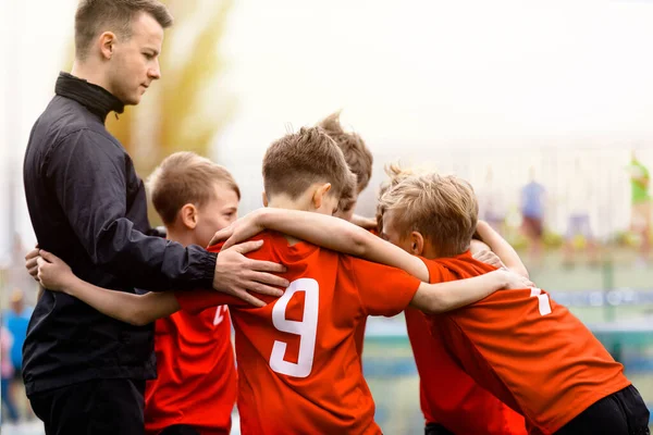 Youth Sports Team. Kids with junior soccer coach huddling in a circle. Boys team in red shirts gathering together to strategize, motivate or celebrate. Football boys achieving tournament success